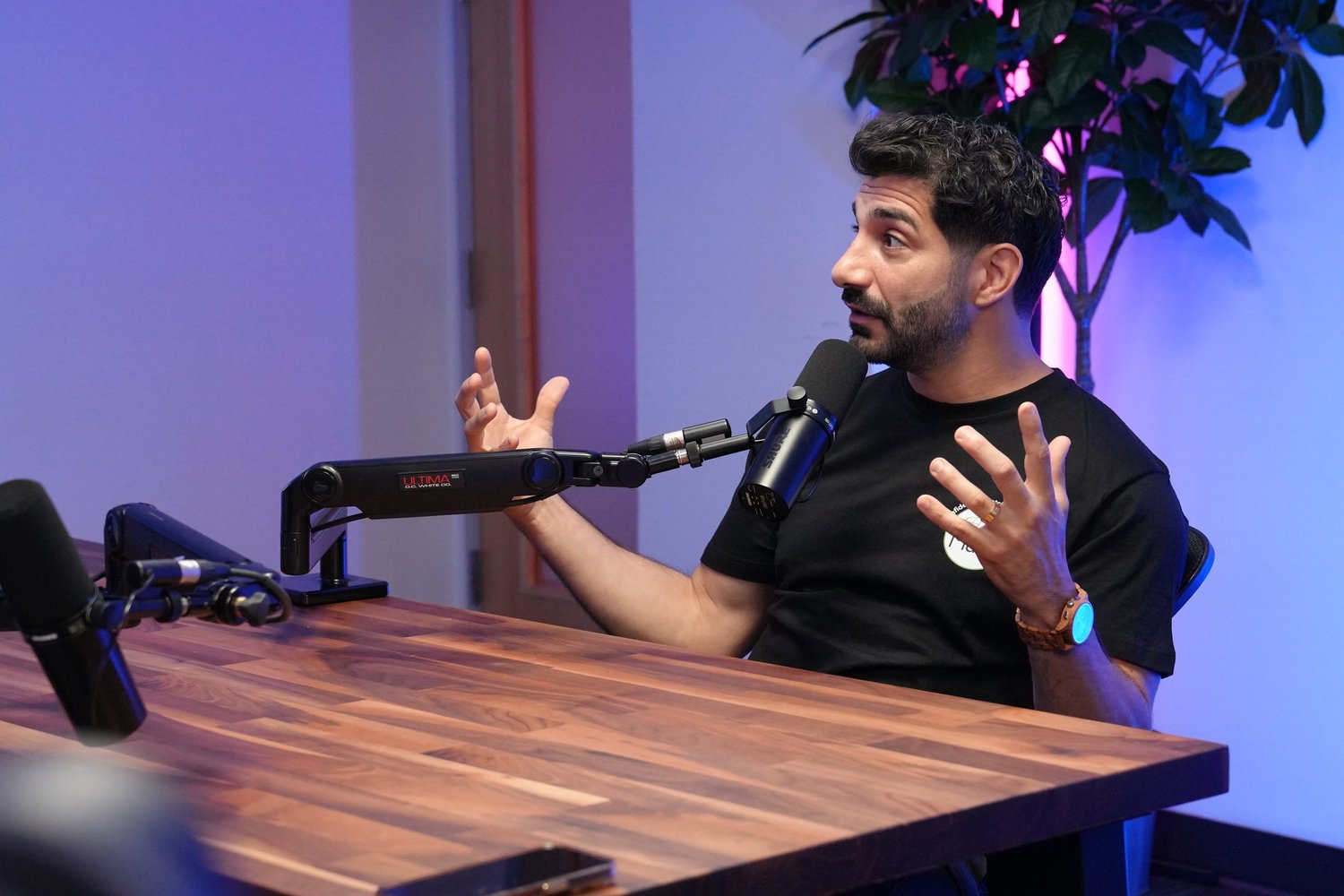 Man at a wooden table with microphones gesturing